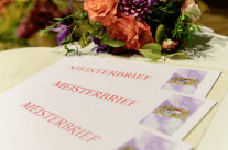 Meisterbriefe liegen überlappend voreinander auf Tisch mit Blumenschmuck