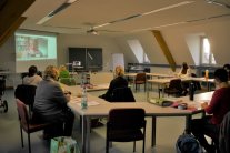 Personen sitzen um Konferenztisch und schauen Richtung Leinwand an einer Wand des Raums