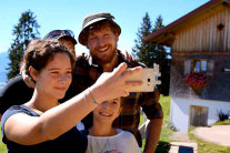 Mutter macht Selfie mit Vater, Tocher und Bergbauern. Im Hintergrund die Alm und Berge.